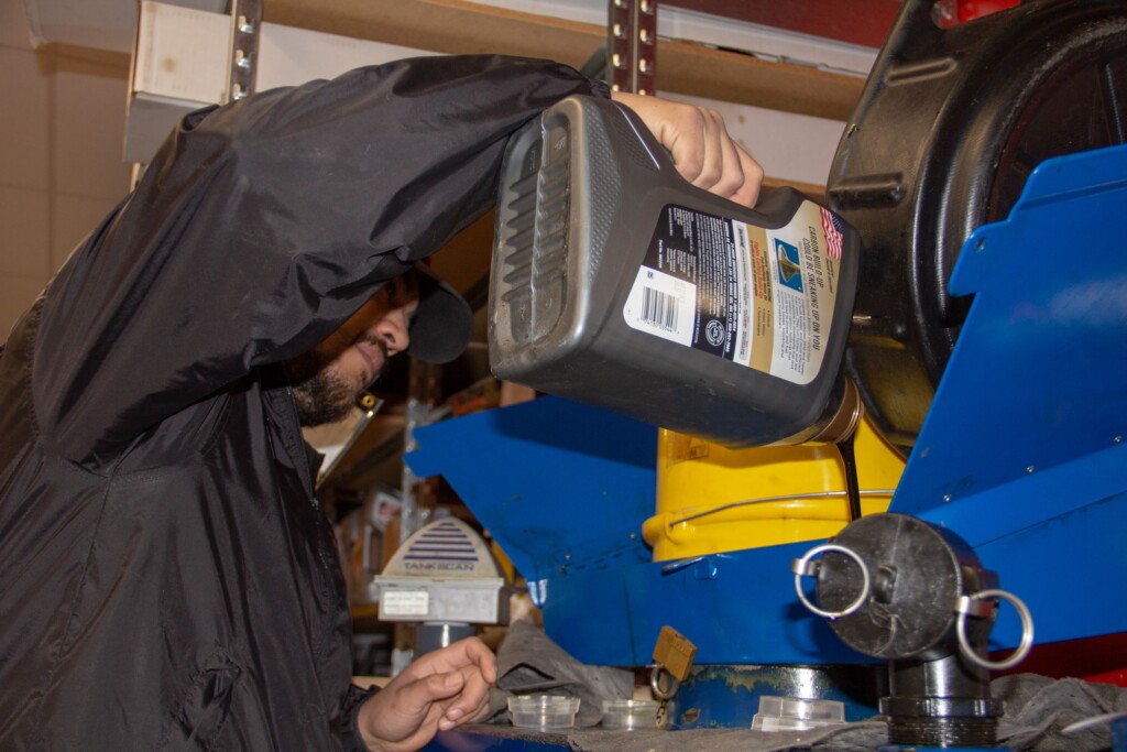 A person in a black jacket pours liquid from a gray container into a blue and yellow machine, likely performing maintenance work in a workshop setting.
