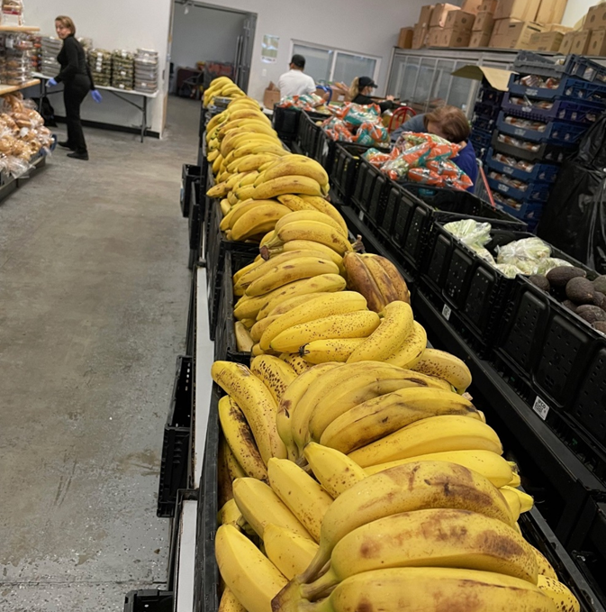 Stacks of ripe bananas fill a long table in a food bank. People gather in the background, collecting food items in a busy, supportive atmosphere.
