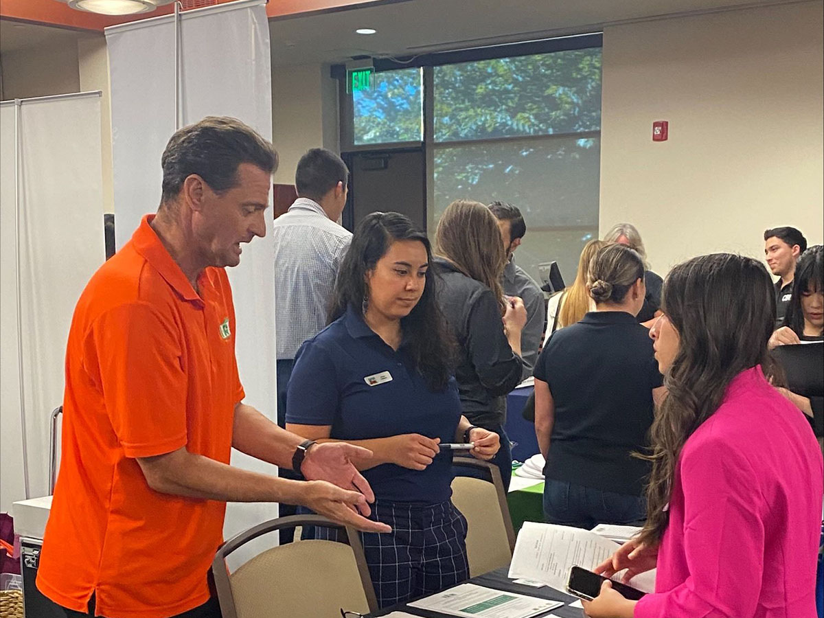 A man in an orange shirt talks to two women at a table during a professional indoor event, with other people engaged in conversation in the background.