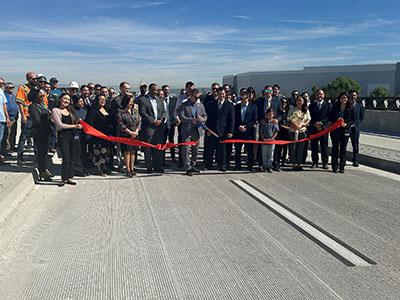 A group of people in formal attire stand on a new road, holding a long red ribbon for a ribbon-cutting ceremony under a clear blue sky.