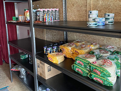 Shelves partially stocked with canned goods, packaged tortillas, and other food items, suggesting a community food pantry setting.