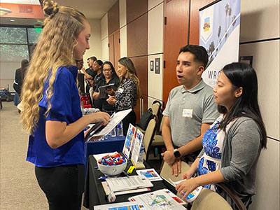 A woman in a blue shirt talks to two people at a booth during a professional event. The atmosphere is engaged and informative, with materials on the table.