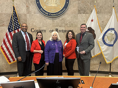 A group of five individuals stands smiling in a formal setting, with the County of Riverside emblem, U.S., California, and county flags in the background.