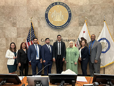 A group of eight professionally dressed individuals stand smiling in a government chamber, with flags and the Riverside County seal behind them.