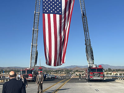 A large American flag hangs between two fire truck ladders on a road with people walking beneath. Clear blue sky and distant mountains in the background. Patriotic tone.