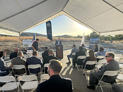 A speaker at a podium addresses an audience seated under a white tent. The setting sun casts warm light over a rural landscape, creating a serene atmosphere.