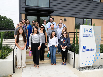 A group of people stands on the steps outside a modern building, smiling. A sign reads 