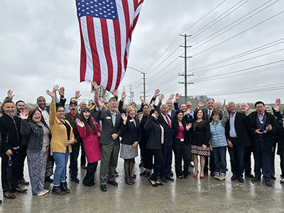A diverse group of people stand outdoors on a rainy day, smiling and waving under a large American flag. The mood is celebratory and unified.