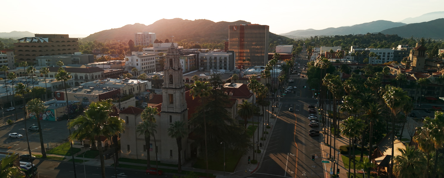 Aerial view of downtown Riverside, California, at sunset, showing palm-lined streets, historic buildings, and the surrounding mountains in the background under a warm golden light.